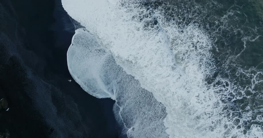 Aerial view of ocean waves breaking against the black beach
