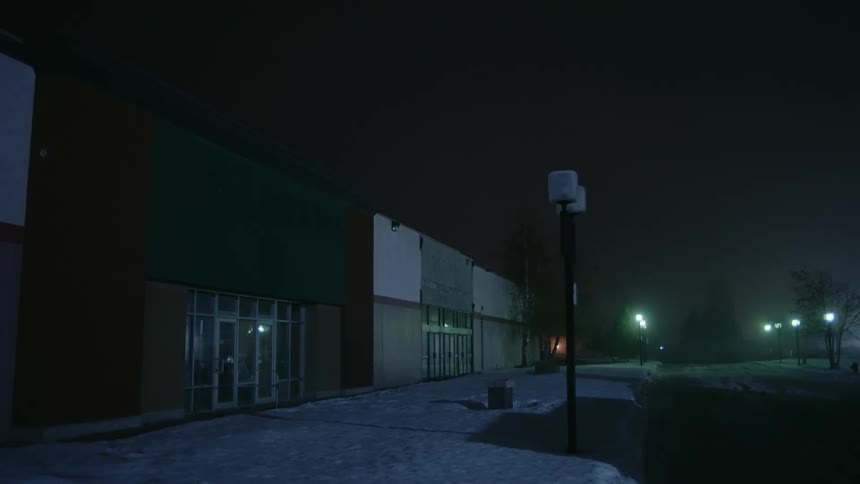 Street lights illuminate the road and a snow covered sidewalk in front of an abandoned building on a foggy winter night