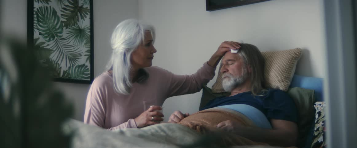 A man is lying in bed sick and his wife is holding a glass and rubbing his forehead with a cloth soaked in cold water