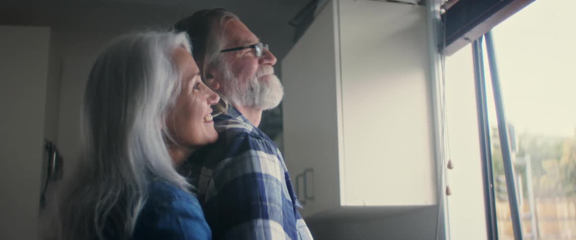 A woman hugs her husband from behind while he looks out a window