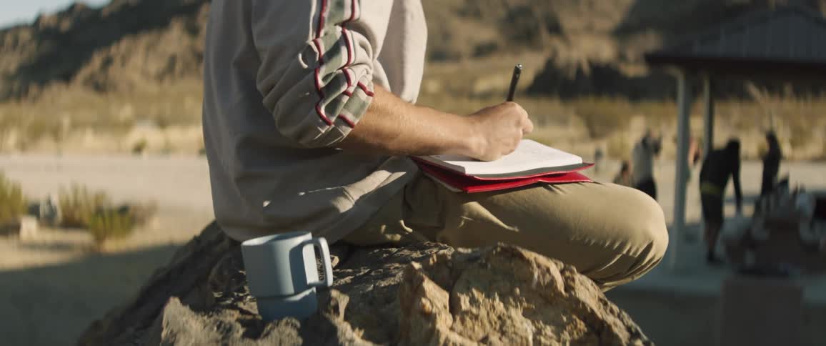 A man sits on a rock and writes in a notebook as others stand by a picnic table