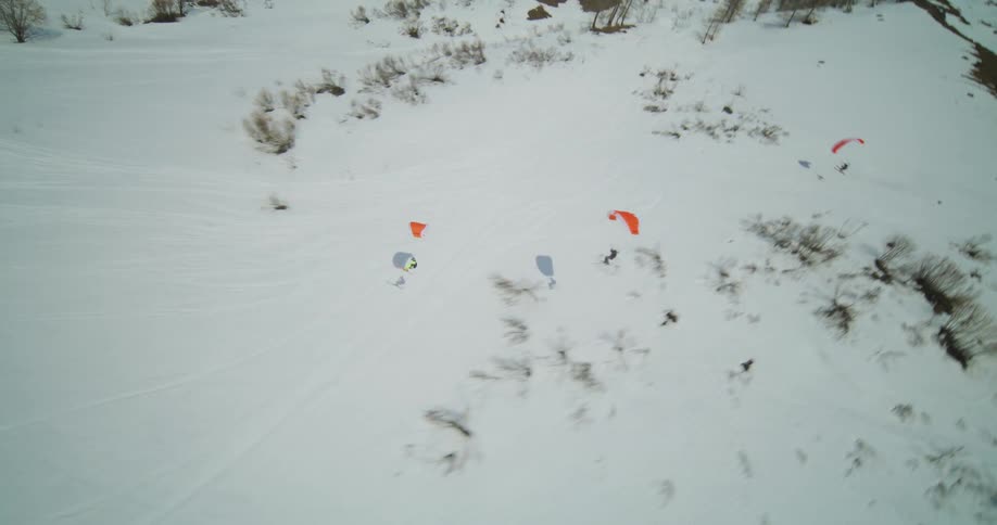 Three people are parachute skiing down a steep snowcovered mountain with a fourth person resting on the snow