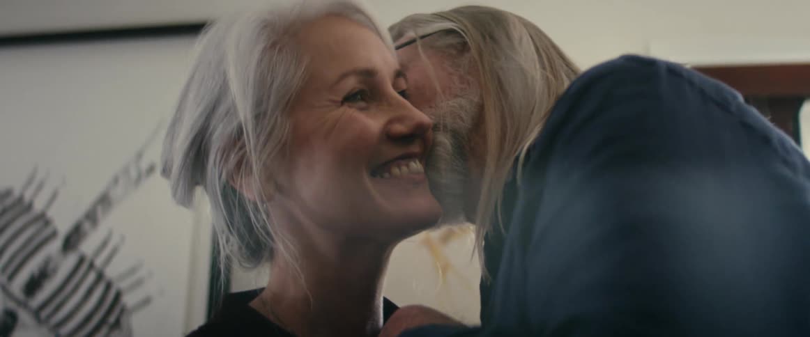Elderly man with long gray hair glasses and beard and woman with gray hair and brown shirt dance happily and kiss