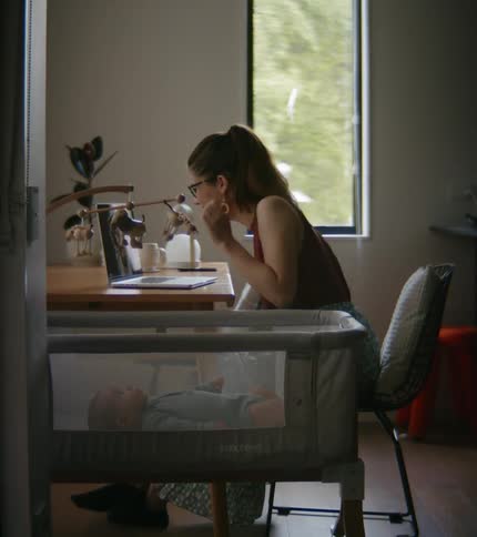 Mother sitting at table makes video call on laptop near the crib