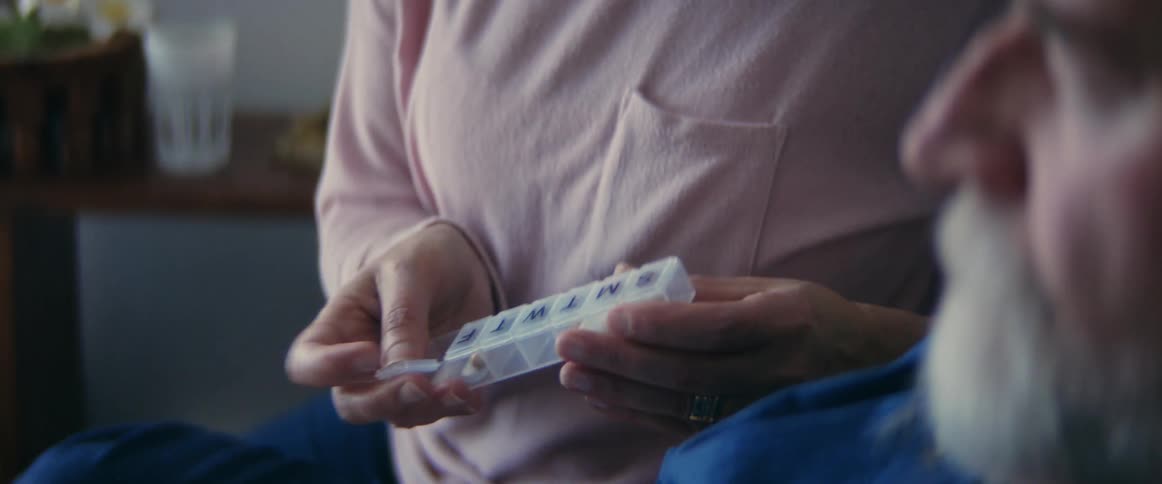 Woman in pink shirt takes pill from daily pill box in order to give elderly man