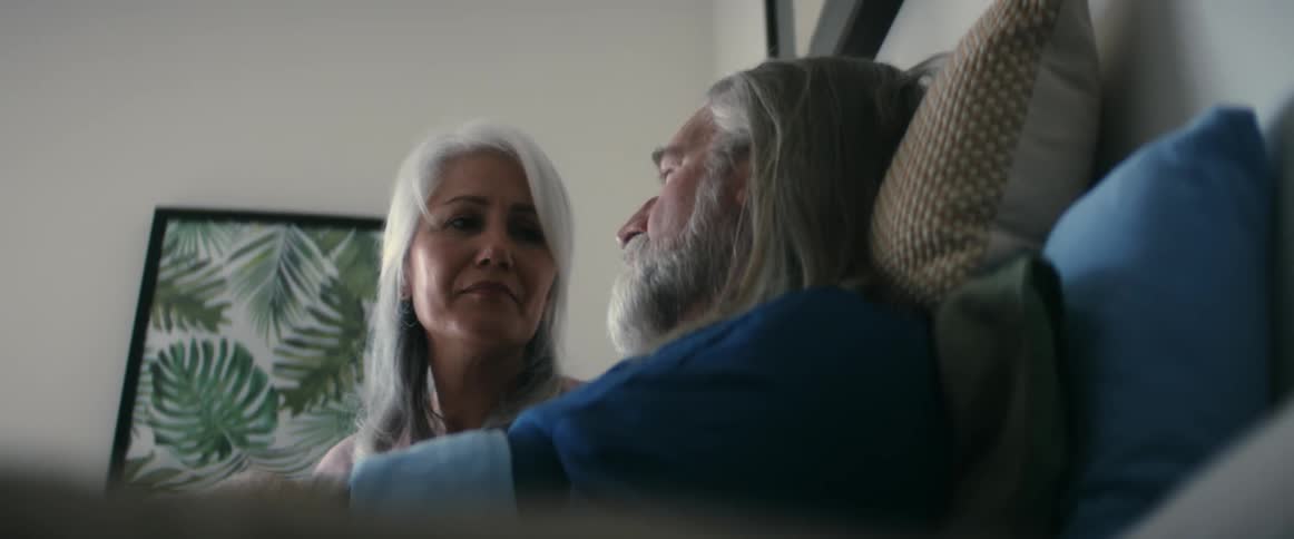 A woman helping a man take pills and drink water while he sits against pillows