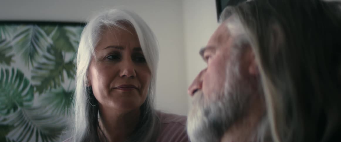 An elderly woman smiles and wipes an elderly mans face as he talks to her