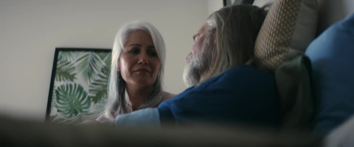 Man with blue shirt long gray hair and beard sits on bed next to woman with gray hair and looking at him with affection