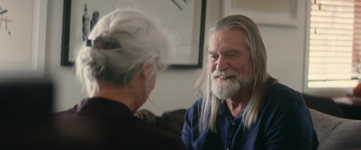Two elderly people laugh together while sitting on a couch in a living room