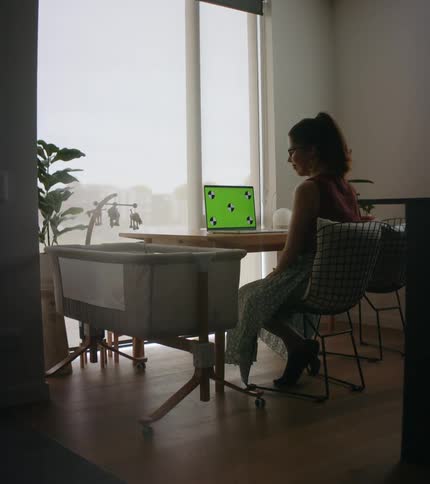 Mother with ponytail and glasses checks on baby in crib while using laptop
