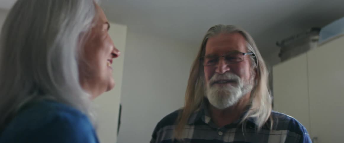 Elderly man with long gray hair glasses and beard cuddles and play with woman with gray hair and blue shirt in the kitchen