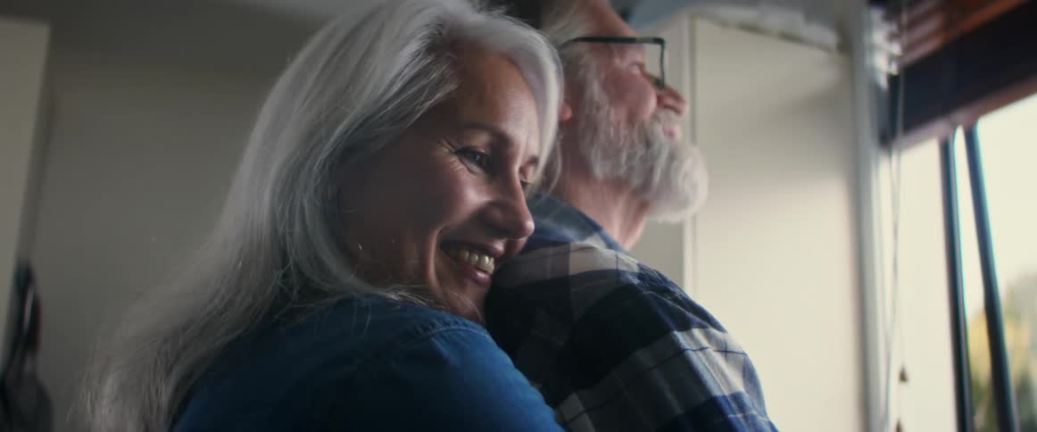 Happy older couple hugging indoors while looking out the window