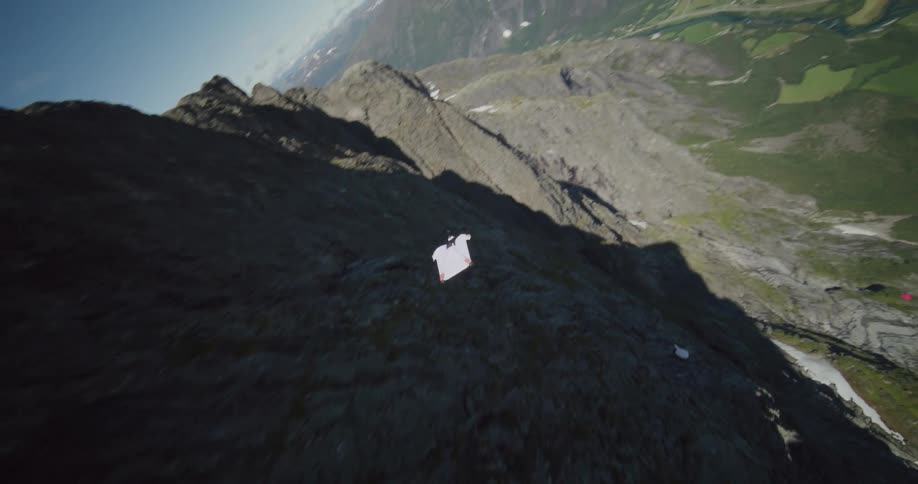 A person wearing a wingsuit is flying along the side of a mountain cliff as they float down to the valley below