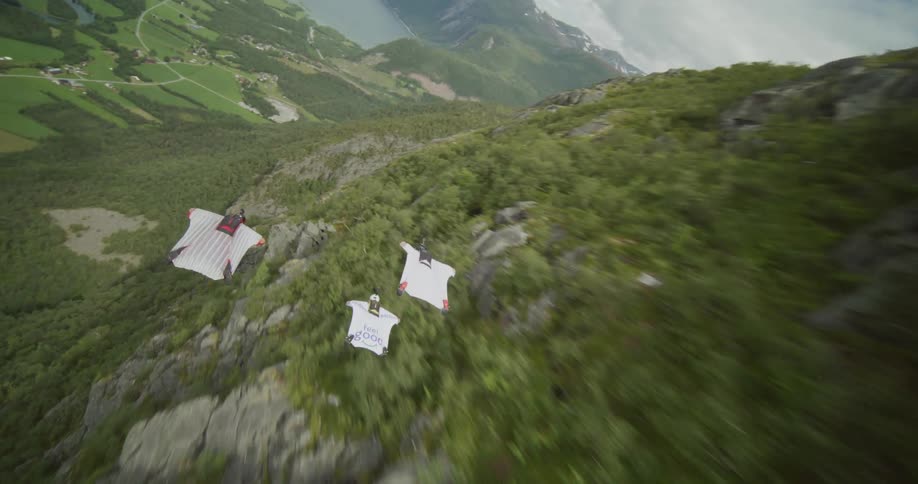 Three people wearing wingsuits are floating along the side of a mountain toward a lake in the valley