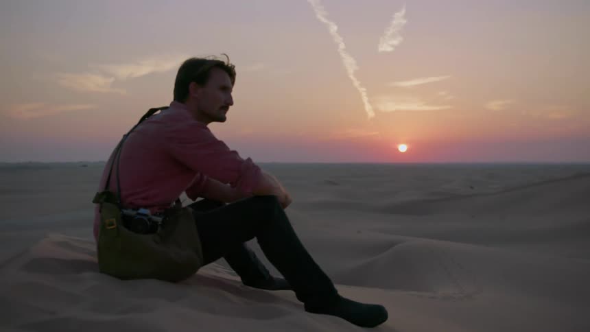 A man sits on a sand dune watching the sun set
