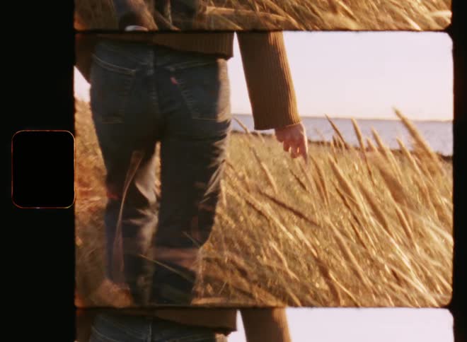 Young woman wearing stylish retro jeans is walking or strolling through a wheat field or grassy area along the coast as her hand is touching the grass plants enjoying the evening sun by the lake or sea beach with relaxed summer vibes