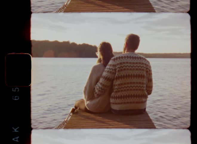 A young man embraces a woman as they sit together on a wooden pier at a lake to watch the sunset during a romantic date night on a warm summer evening on vintage Super 8 mm film