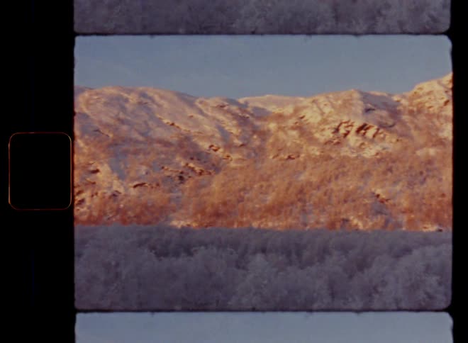 Panning Super 8 panorama wide shot of a snowy landscape in winter wonderland with a snow-covered valley with a forest with bare trees and a high mountain glowing in the warm sun shine as the sun sets in the evening time during golden hour on a clear day