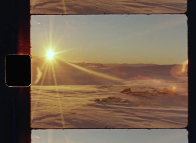 Old retro Super-8 footage of a rocky plain covered in snow and ice as the sun rises at the horizon behind the mountain ridge brightening and shining into the valley on a freezing cold winter morning above the arctic circle in unspoiled pristine nature