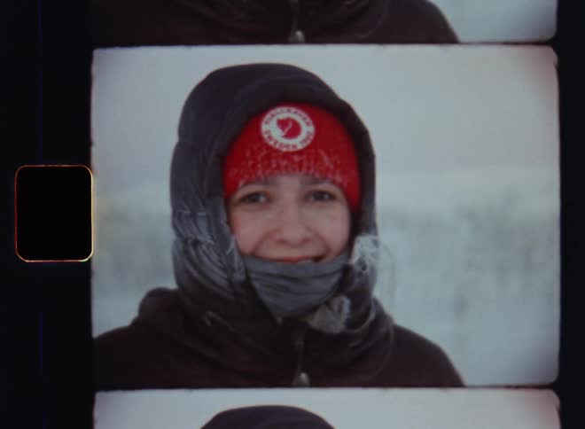 Old super 8mm film footage of a beautiful young woman wearing a red beanie bundled up in a black coat with hood and scarf smiling at the camera while her hair is frozen in the blizzard by the harsh weather and force of Mother Nature in the mountains