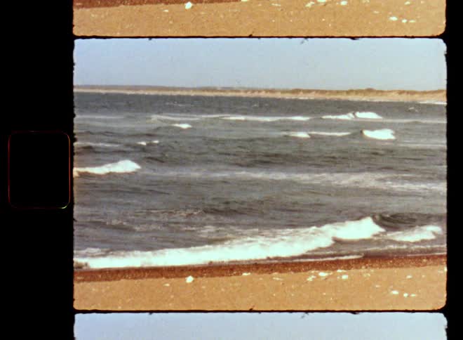 Old film footage of an ocean or sea bay while breaking waves with white foam caps are crashing onto the sandy beach on a sunny day with blue sky at the coast