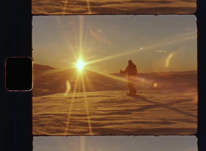 A man with snow shoes or hiking skis and trekking poles walking across snow-covered ground of a mountain plateau into the setting evening sun on a cold day during arctic Scandinavian winter on vintage 8mm film with a nostalgic vibe and lens flare