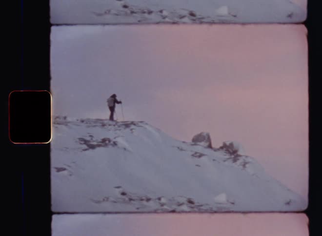 Handheld 8mm film of a person wearing outdoor clothes and a backpack hiking or skiing on a snowy and rocky mountain ridge as a silhouette against the glowing evening sky during sunset on a freezing cold winter day after a blizzard with snowfall