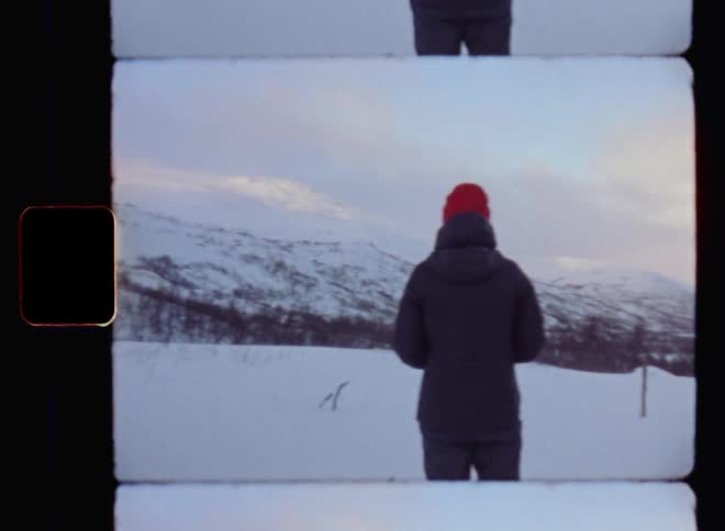 Handheld 8mm film footage of an unrecognizable young woman outdoors with a black winter jacket coat and red hat contemplating while standing and watching the snow-covered peaks of a high mountain range as the clouds are hit by warm sunshine