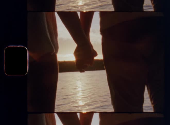 Young man and woman holding hands facing a body of water as the sun sets during their first romantic date on a warm spring evening on old 8mm film