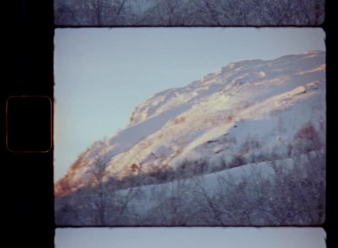 Handheld shaky 8mm film footage of a mountain face with steep cliffs and rocky slope with bare trees covered in snow and ice on a sunny but extremely cold winter day with frosty temperatures well below freezing during sunrise in early morning sun