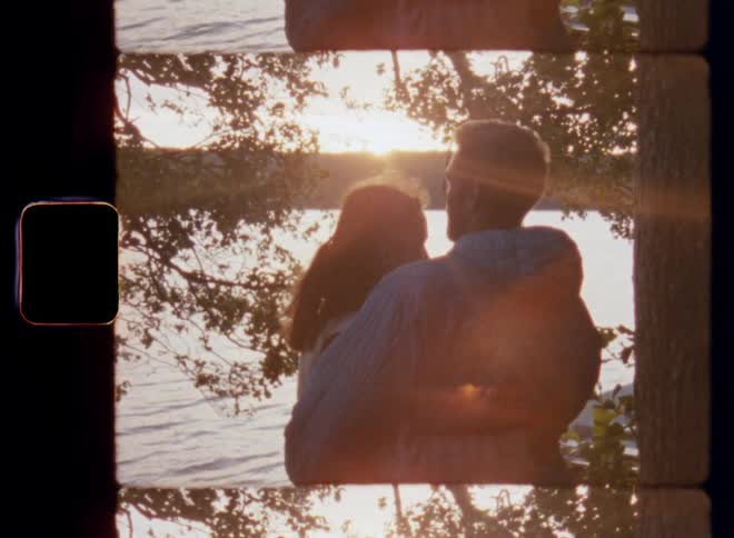 Super 8mm film of a Caucasian loving couple consisting of man and woman standing under a tree and hugging each other as they watch the sunset over a forest lake and the sun reflects on the water
