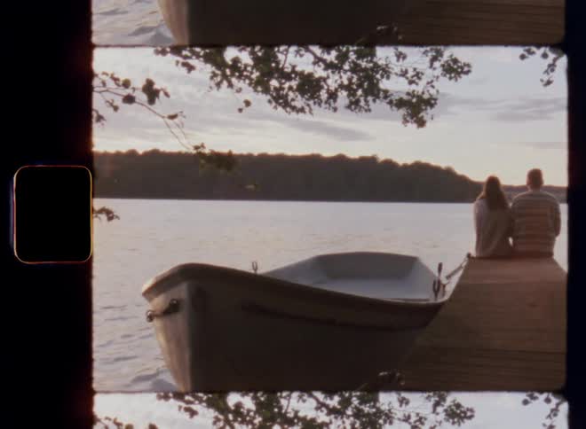 Two friends consisting of a Caucasian man and young woman are sitting on a wooden bridge next to a rowing boat looking at the water as the sun sets and the last sun rays flare through the branches of the trees surrounding the bay of the forest lake
