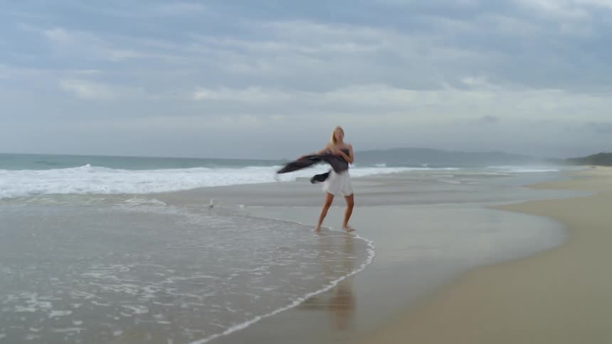 Blonde woman holds wrap to cover her chest while walking along beach on windy day