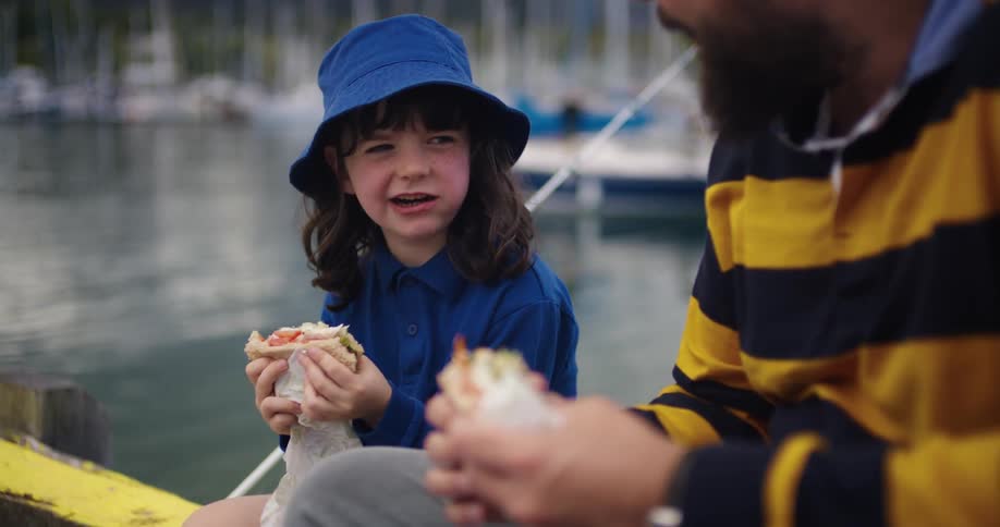Father and daughter sitting on a peer near the water enjoying sandwiches and talking