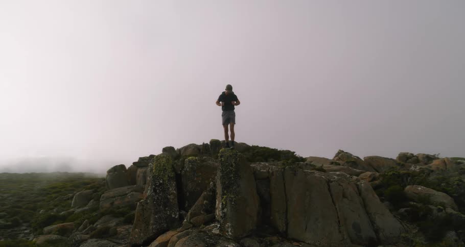 Man with cap shirt shorts and backpack stands on the edge of large rock in the field on a cloudy day