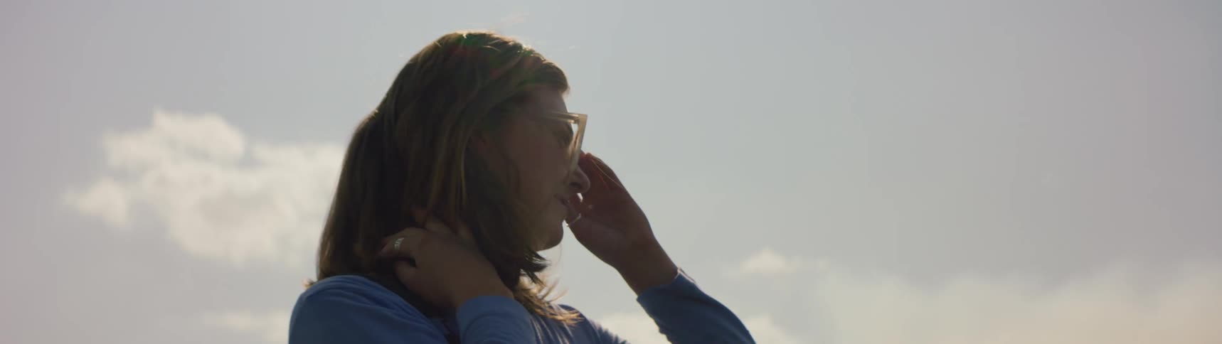 Woman with brown hair blue shirt touches her sunglasses under shining sun smiling