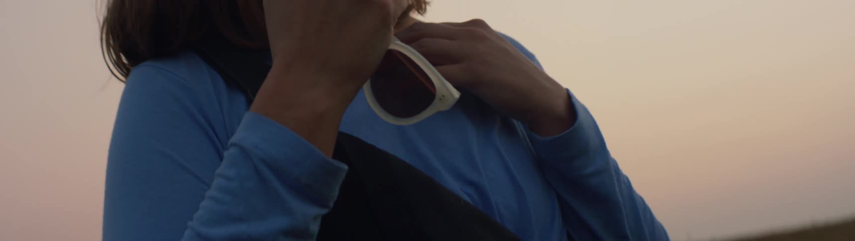 Woman wearing blue clothes removes her sunglasses from her head while gazing at the sunset