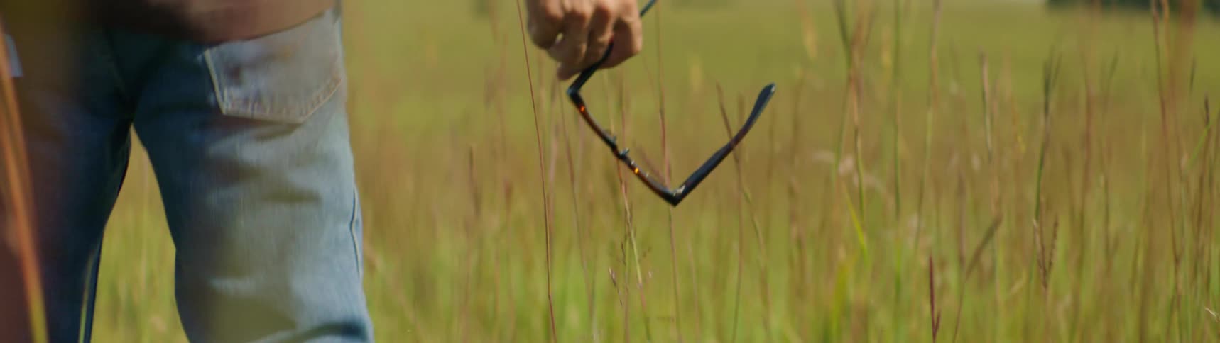 A man is carrying his glasses in his hand as he walks in through weeds in a field