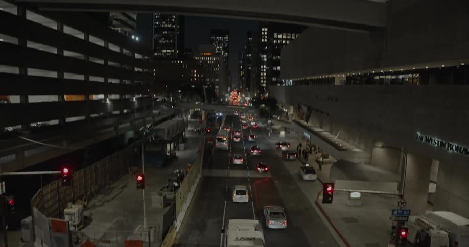 Traffic is moving on a road into downtown as people walk on the sidewalks and lights glow on tall buildings and towers in the night sky