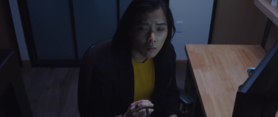 A woman is sitting at a desk with a computer as she plays with her pen in her hand and looks around before looking at the computer monitor