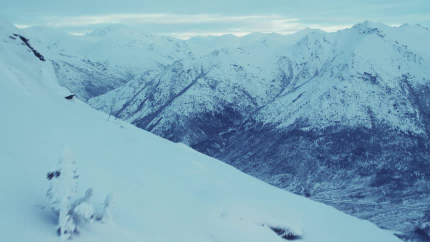 Moving over a snow covered mountain there are mountains in the distance and a valley below