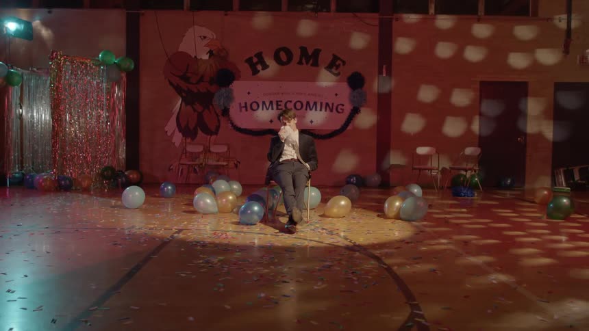 A boy sitting in the middle of a gymnasium decorated with balloons and confetti with a disco light going around