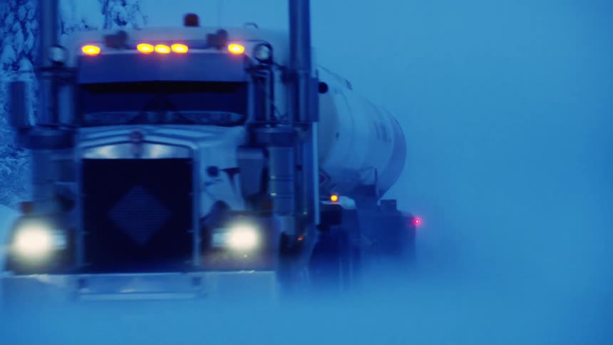 A tanker truck is traveling through a blizzard on a road lined with snow covered trees