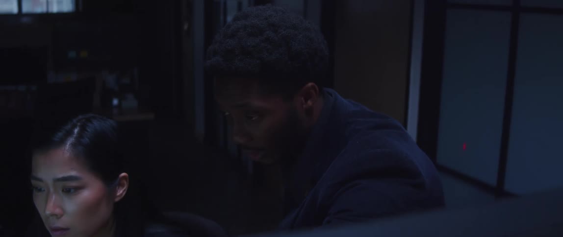 A woman is sitting at a desk looking at a computer monitor as a man looking over her shoulder talks to her