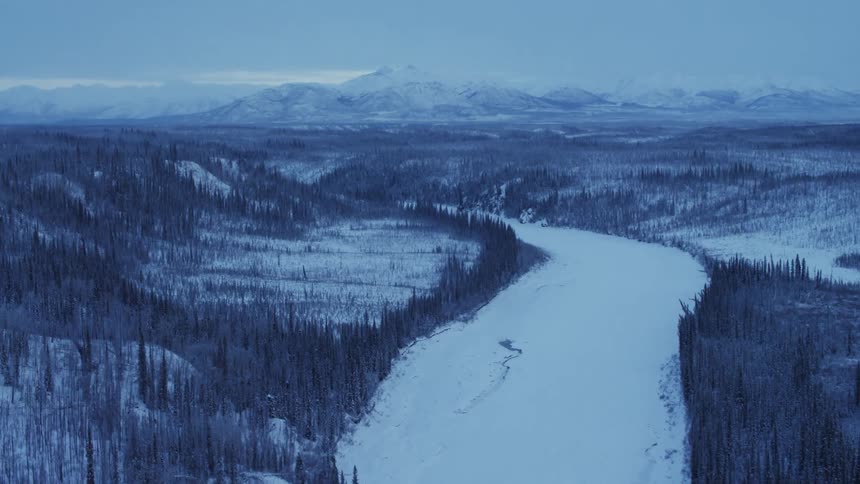 A thick blanket of snow covers a river that runs through a forest near a mountain range