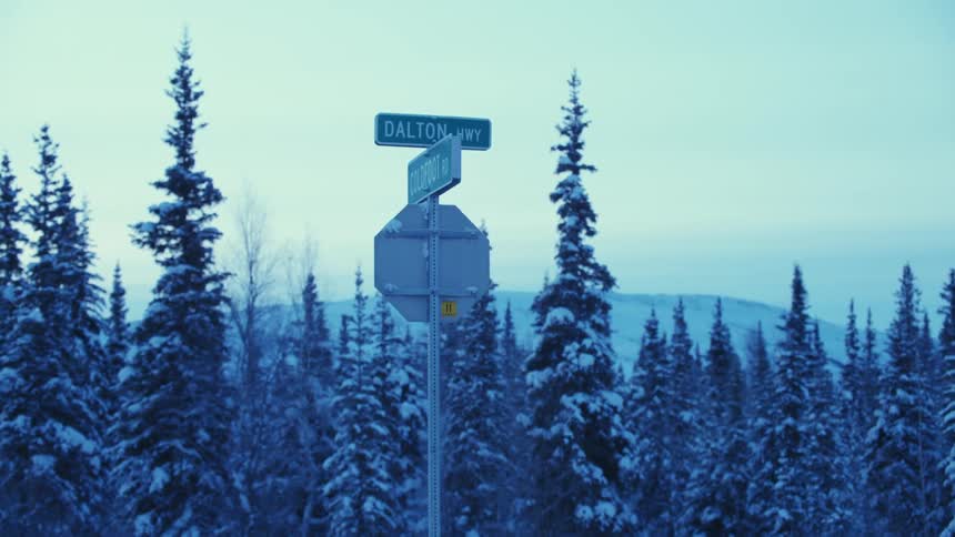 Road signs stand at the corner of coldfoot road and the dalton highway with snow covered trees and mountains in the distance on a winter day