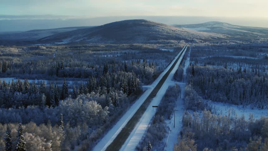 An aerial view of a long paved road passing through a forest of trees during winter