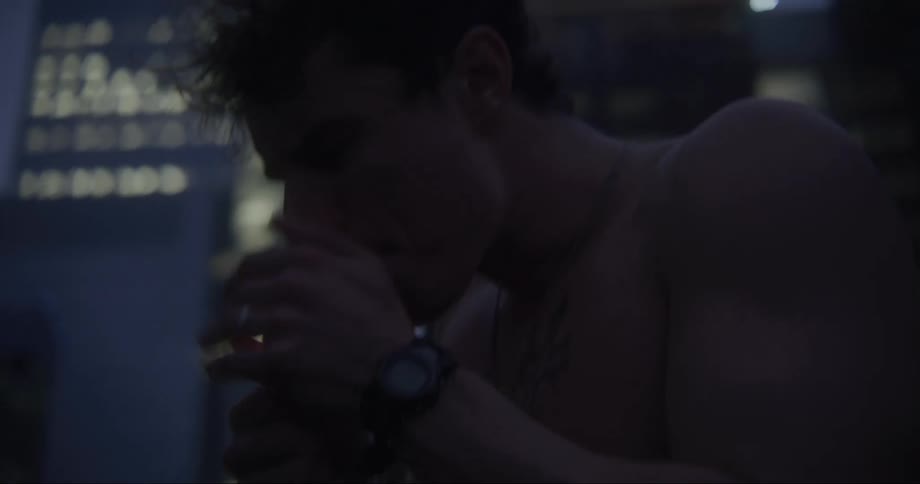 A bare chested teenage boy is standing outside lighting a cigarette as lights in tall buildings glow in the distance