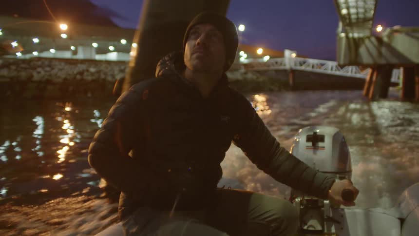 A man wearing a black cap and coat driving a motor boat through a river under a bridge