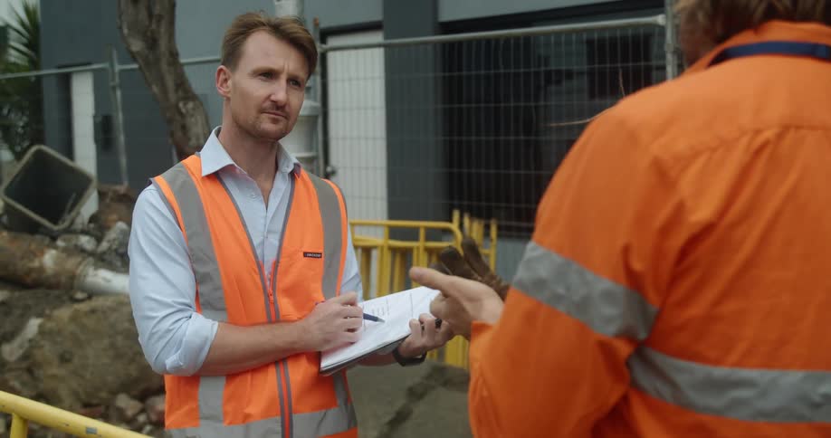 A man with a vest a pen and a book standing in a construction site talking to another man with a fence and a building in the background